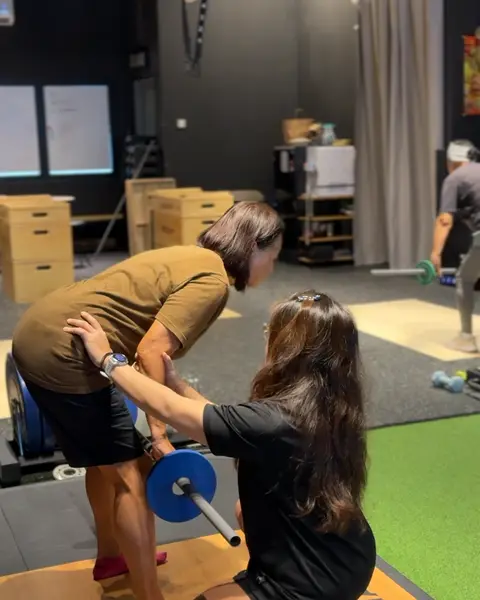 A coach with one hand on an older member's back, guiding him through a hip hinge with a light barbell.
