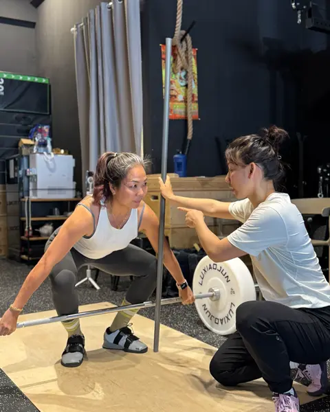 A coach crouching beside a member in a deep squat, checking her position against a vertical pole.