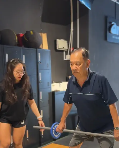 An older member holding a light barbell at arm's length while a coach watches his setup.