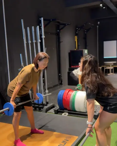 An older member lifting a light barbell from the floor as a coach guides her technique.