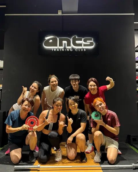 A strength and conditioning class posing with plates and dumbbells under the ANTS Training Club sign.